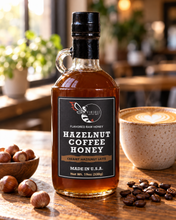Load image into Gallery viewer, Firebee Hazelnut Coffee Honey 19oz glass bottle on a cafe table with a latte, hazelnuts and coffee beans